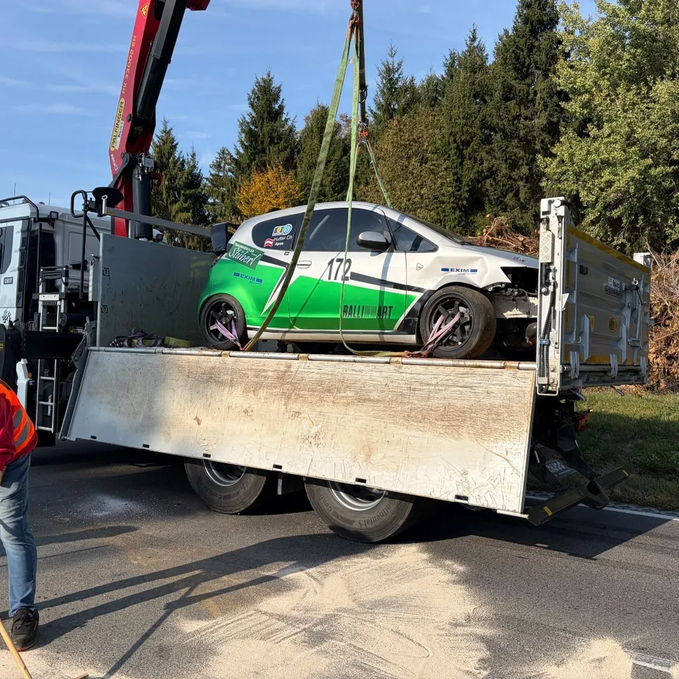 Ein Kran hebt ein beschädigtes grünes und weißes Auto auf einen Tieflader, der am Straßenrand steht.