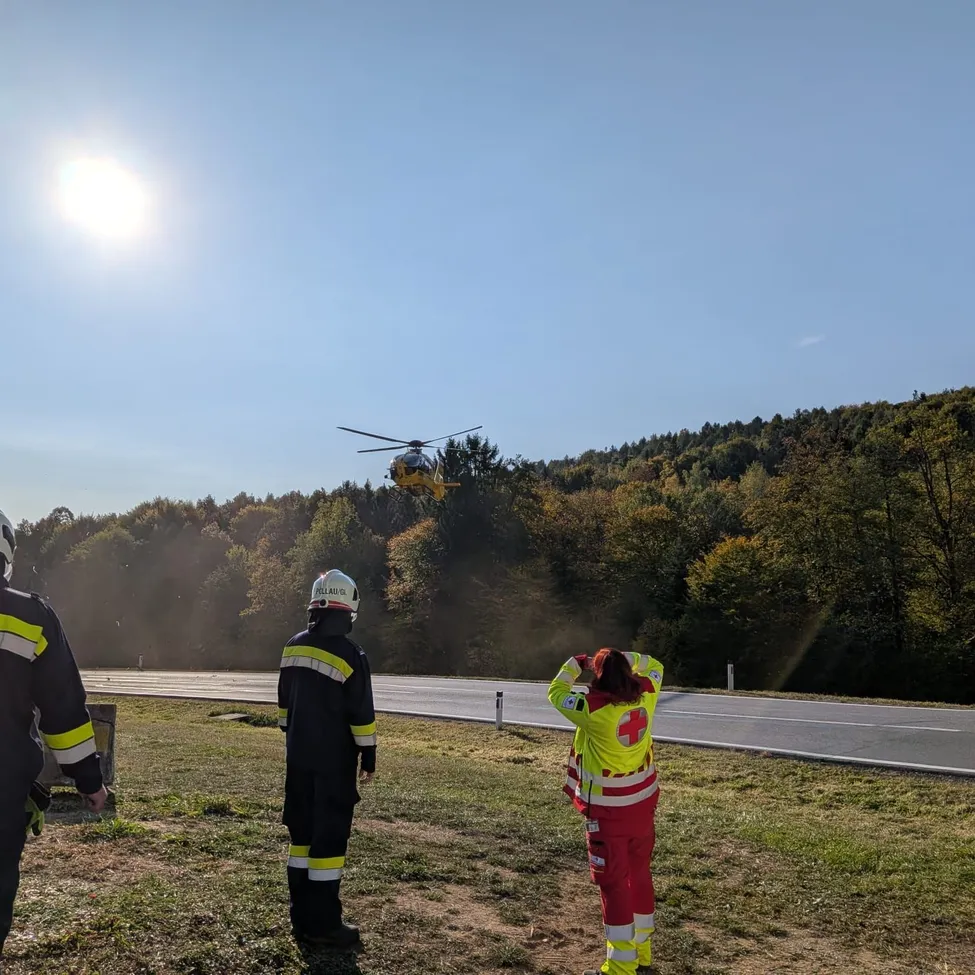 Drei Personen in Sicherheitsausrüstung stehen auf einem Grasfeld und beobachten einen Hubschrauber, der über einer Straße fliegt.