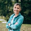 Eine Frau mit blonden Haaren steht mit verschränkten Armen in einem Park. Sie lächelt und trägt eine Jeansjacke.