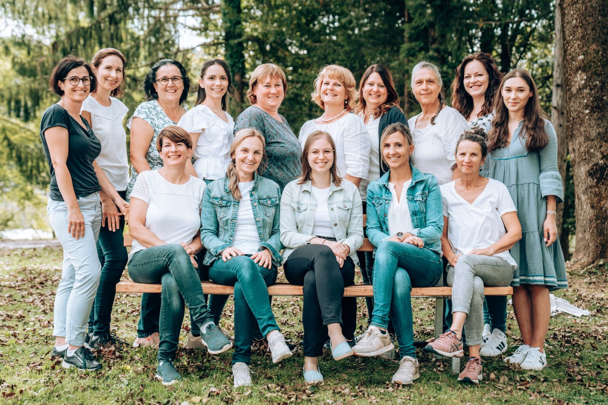Eine Gruppe von Frauen in weißen Oberteilen und Jeans, die für ein Foto auf einer Holzbank in einem Park mit Bäumen im Hintergrund posieren.