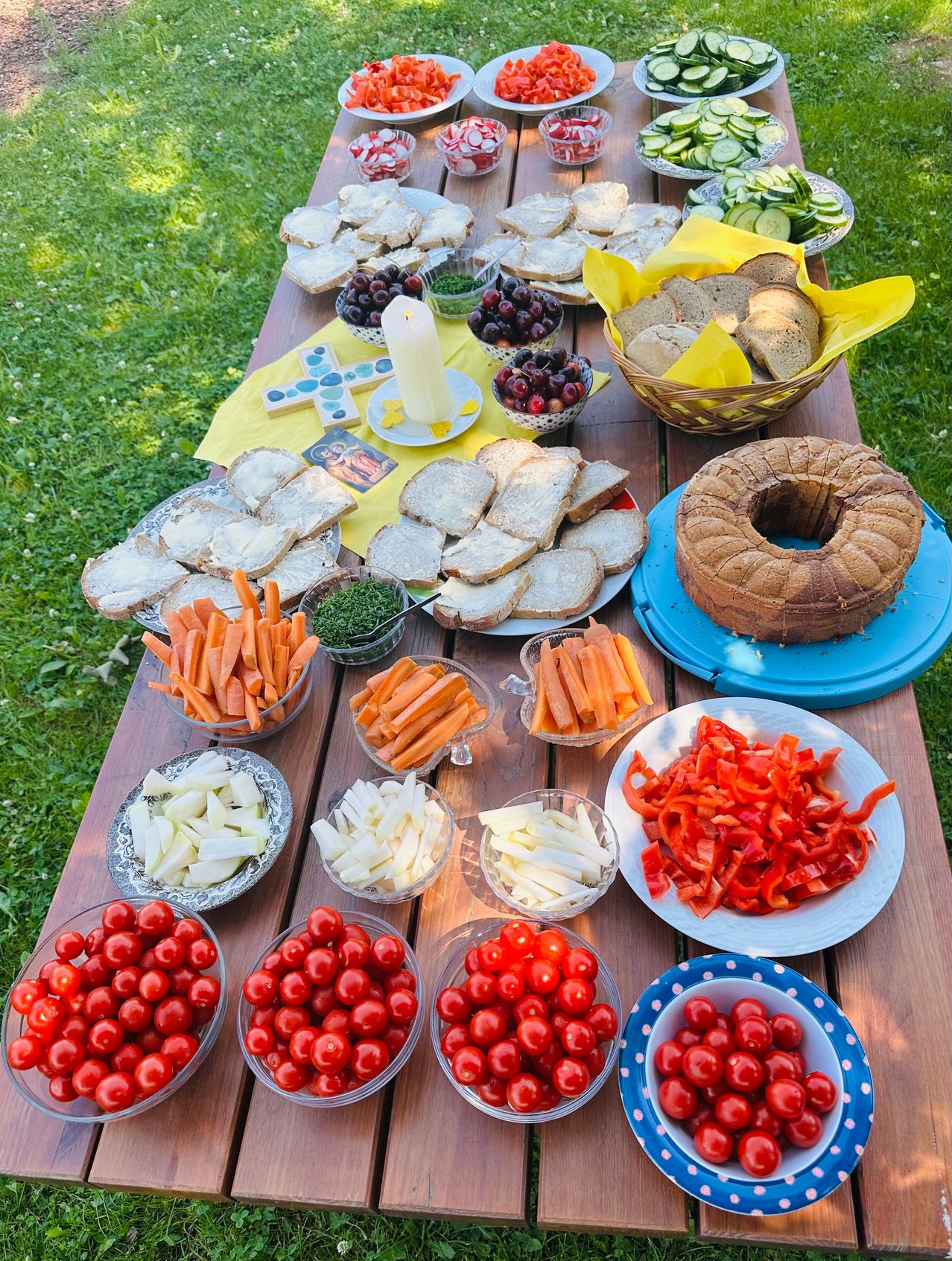 Ein Holztisch im Freien ist mit verschiedenen Speisen gefüllt. Brot, Karotten und rote Paprika sind auf Tellern angeordnet. Tomaten sind in Schalen und ein Guglhupf steht auf einem blauen Teller.