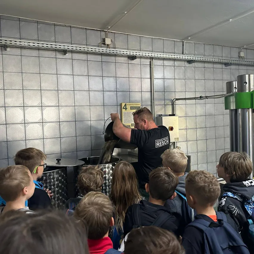 Ein Mann gießt eine Flüssigkeit in einen großen Metallbehälter, während eine Gruppe von Kindern zusieht.