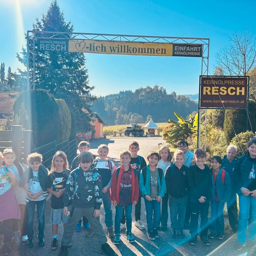 Eine Gruppe von Kindern und Erwachsenen steht vor einem Schild, das 'Kernölpresse Resch' und '-lich willkommen' besagt. Sie stehen vor einem Gebäude mit einem Baum und einem Feld im Hintergrund.