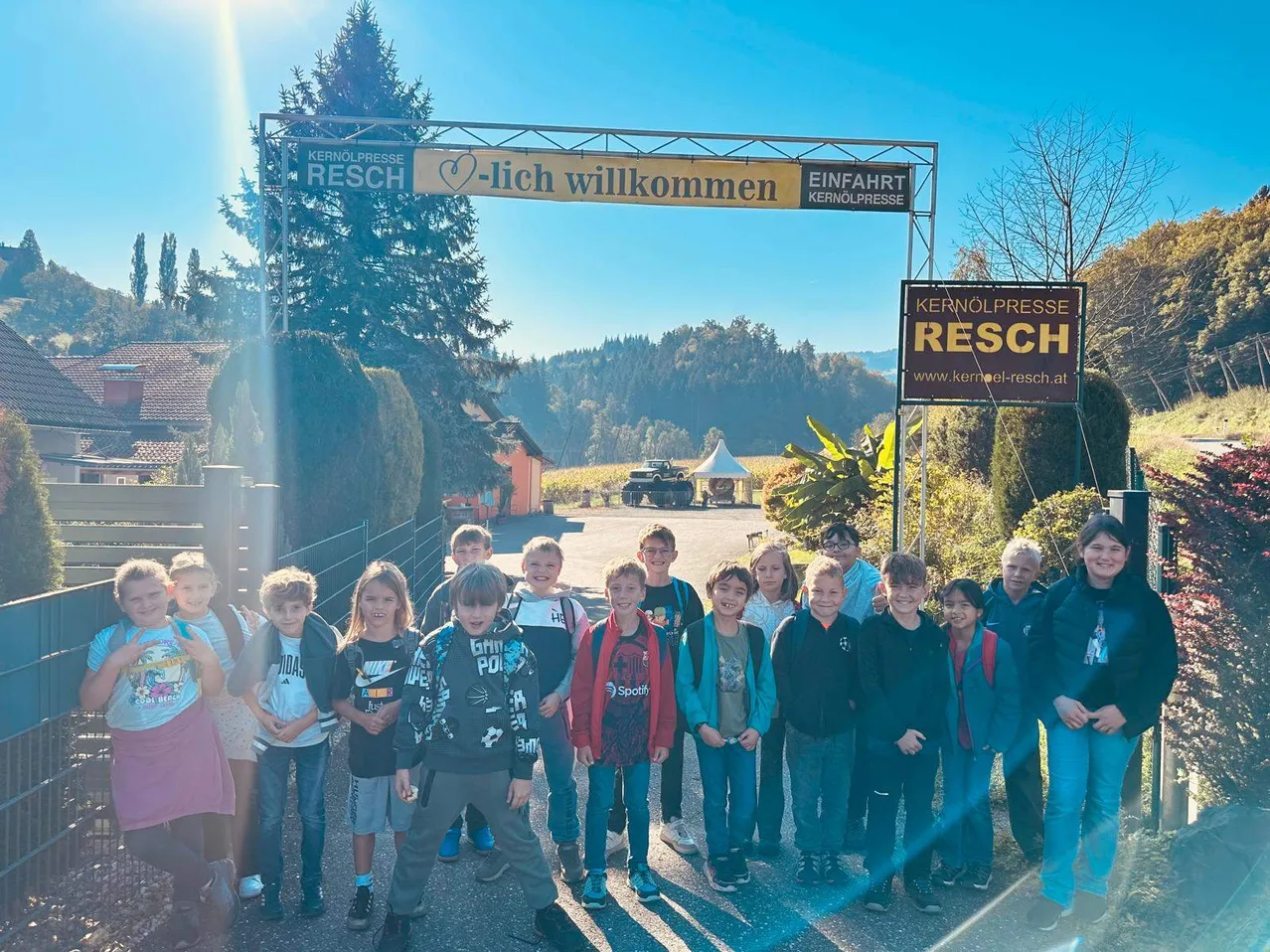 Eine Gruppe von Kindern und Erwachsenen steht vor einem Schild, das 'Kernölpresse Resch' und '-lich willkommen' besagt. Sie stehen vor einem Gebäude mit einem Baum und einem Feld im Hintergrund.