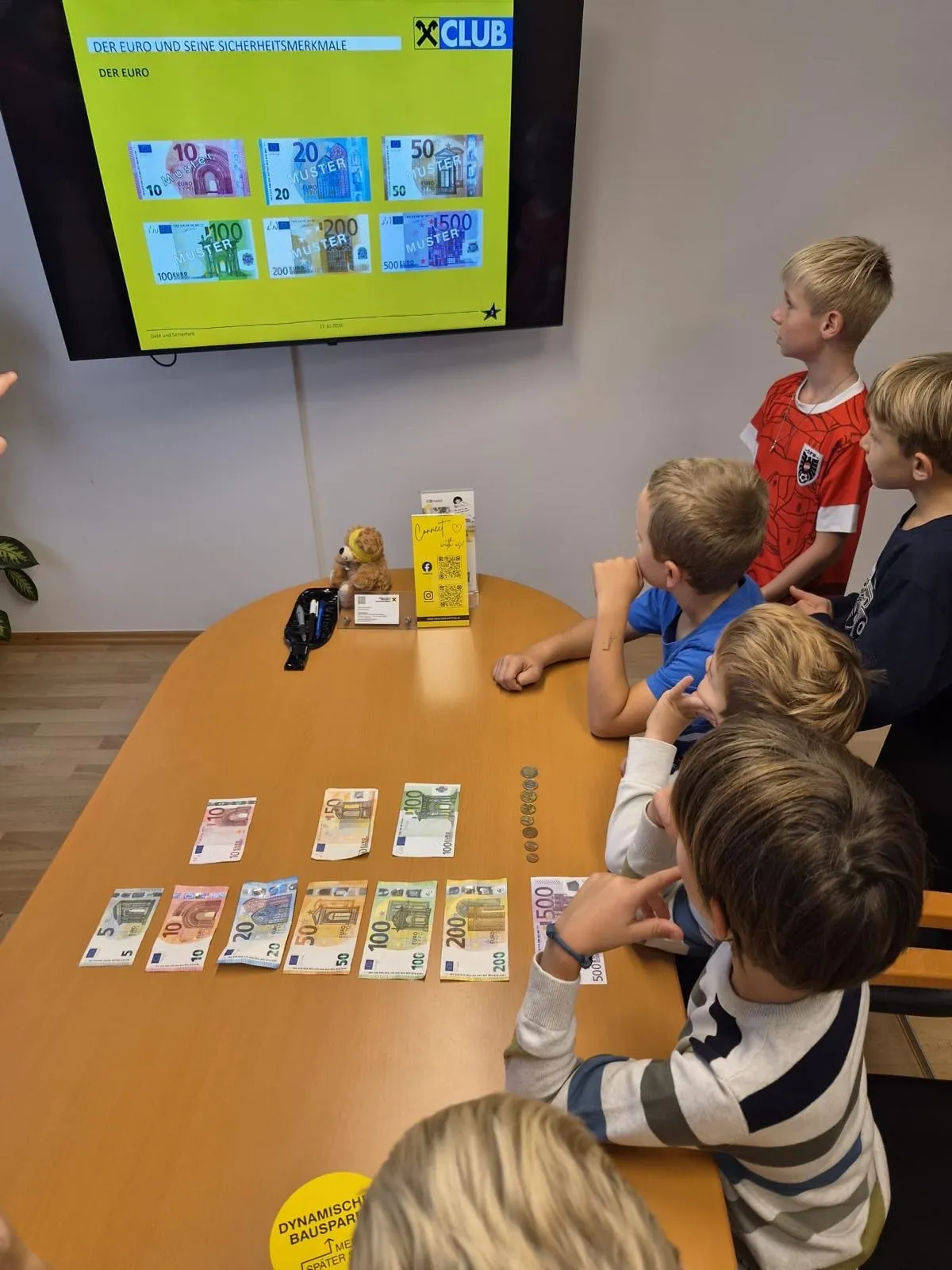 Eine Gruppe von Kindern sitzt um einen Tisch herum mit verschiedenen Euro-Banknoten und Münzen. Ein Monitor ist an der Wand hinter ihnen montiert und zeigt Informationen über Euro.