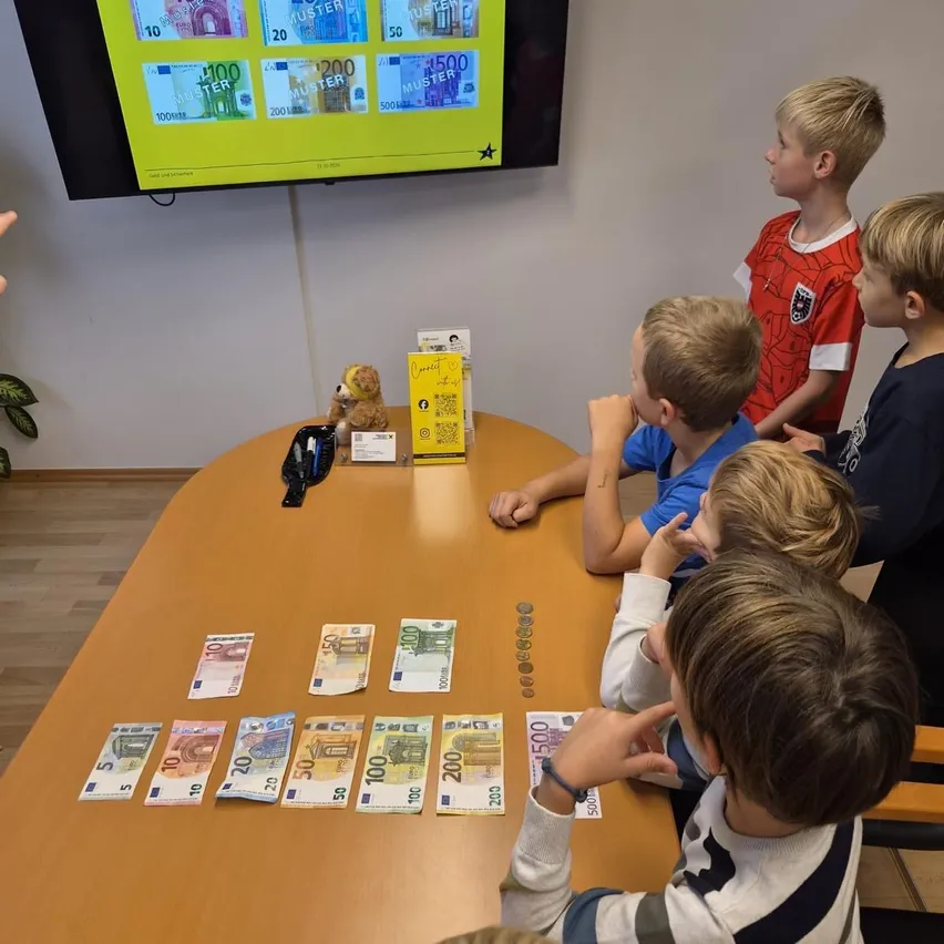 Eine Gruppe von Kindern sitzt um einen Tisch herum mit verschiedenen Euro-Banknoten und Münzen. Ein Monitor ist an der Wand hinter ihnen montiert und zeigt Informationen über Euro.