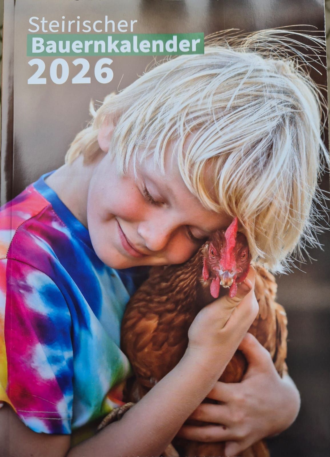 Ein Kind in einem bunten Tie-Dye-Shirt umarmt ein braune Huhn nah an seinem Gesicht, mit dem Jahr 2026 in der oberen linken Ecke.