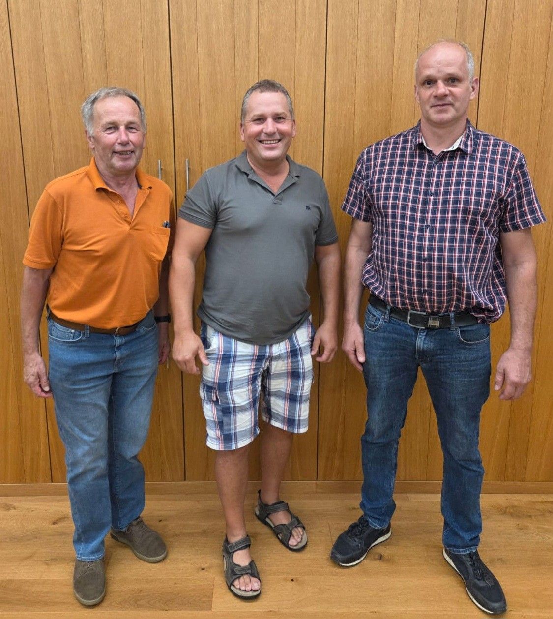 Three men standing next to each other in front of a wooden wall, all smiling and dressed casually.