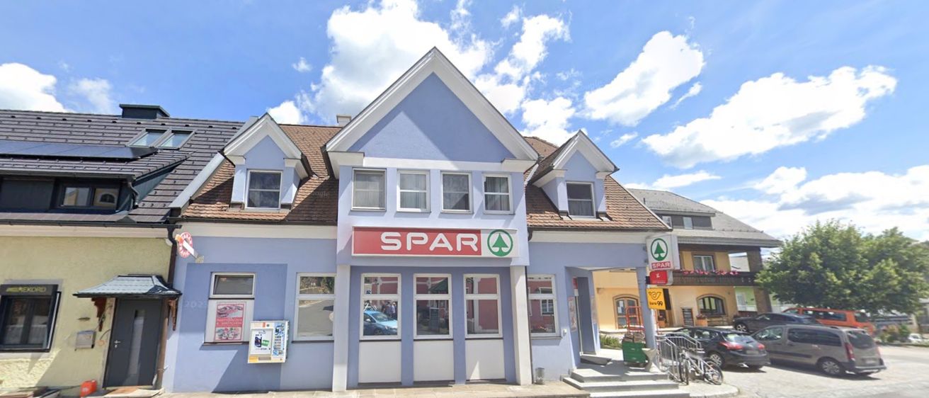 Ein blaues Gebäude mit einem roten Schild, das SPAR liest, mit mehreren Fenstern und einem braunen Dach, befindet sich unter einem blauen Himmel mit Wolken.