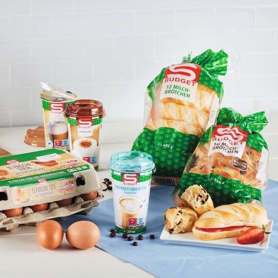 Eine Auswahl an Frühstücksartikeln, einschließlich Brot, Eier und Kaffee, ist auf einem blauen Tuch angeordnet. Das Brot ist mit Budget gekennzeichnet, und es gibt Kaffeepads und eine Tasse Kaffee.