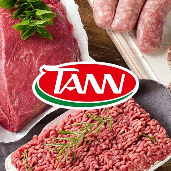 Rohes Rindfleisch und Hackfleisch mit Kräutern und Würsten auf einem Holztisch. TAN-Logo in Rot und Grün.