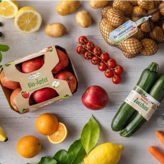 Eine Auswahl an Obst und Gemüse auf einer weißen Oberfläche. Ein Apfelkarton, Orangen, Zitronen, Kartoffeln und Tomaten sind vorhanden.