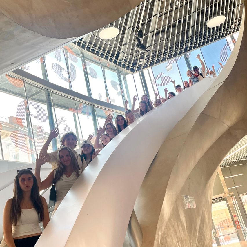 Eine Gruppe von Menschen, darunter Frauen und Männer, steht auf einer gebogenen Treppe in einem modernen Gebäude und hebt die Hände. Glaswände reflektieren den Himmel.