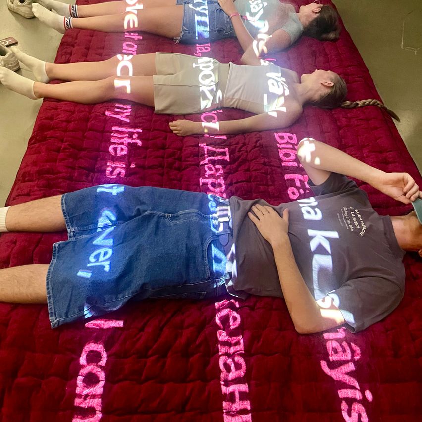 Drei Menschen liegen auf einem roten Bett mit verschiedenen Wörtern darauf. Die Person auf der linken Seite hat ihren Kopf auf dem Kissen. Die Person in der Mitte hat ihre Hände auf dem Bauch. Die Person auf der rechten Seite hat ihre Hand am Kinn.