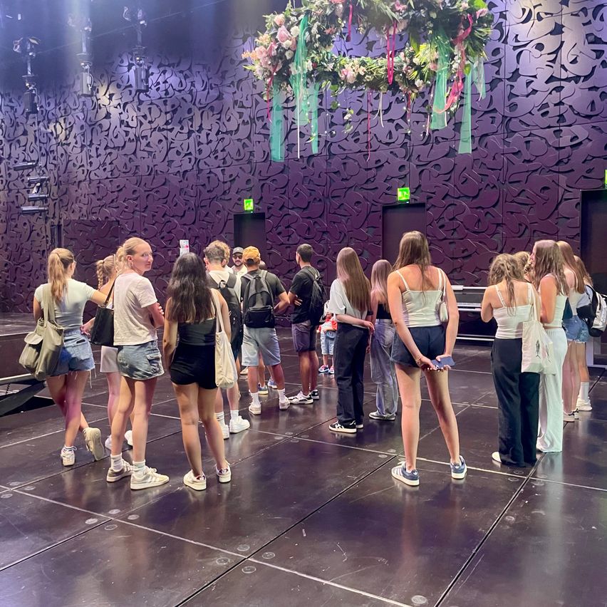 Eine Gruppe von Menschen steht drinnen, einige tragen Rucksäcke, andere Handtaschen. Sie scheinen in einer Schlange zu warten, und es gibt eine dekorative Blumenarrangement darüber.