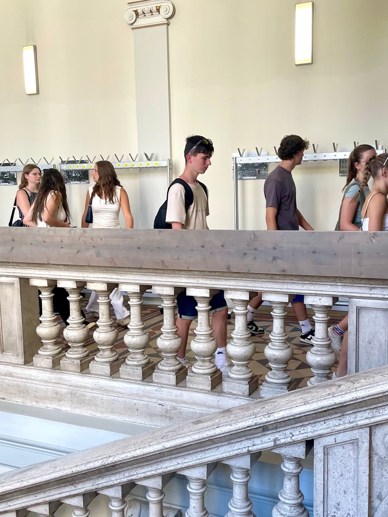 Mehrere Menschen sind auf einem Balkon versammelt. Einige stehen, andere sitzen. Sie sind lässig gekleidet und scheinen Touristen zu sein. Der Balkon hat einen gefliesten Boden und ein weißes Geländer.