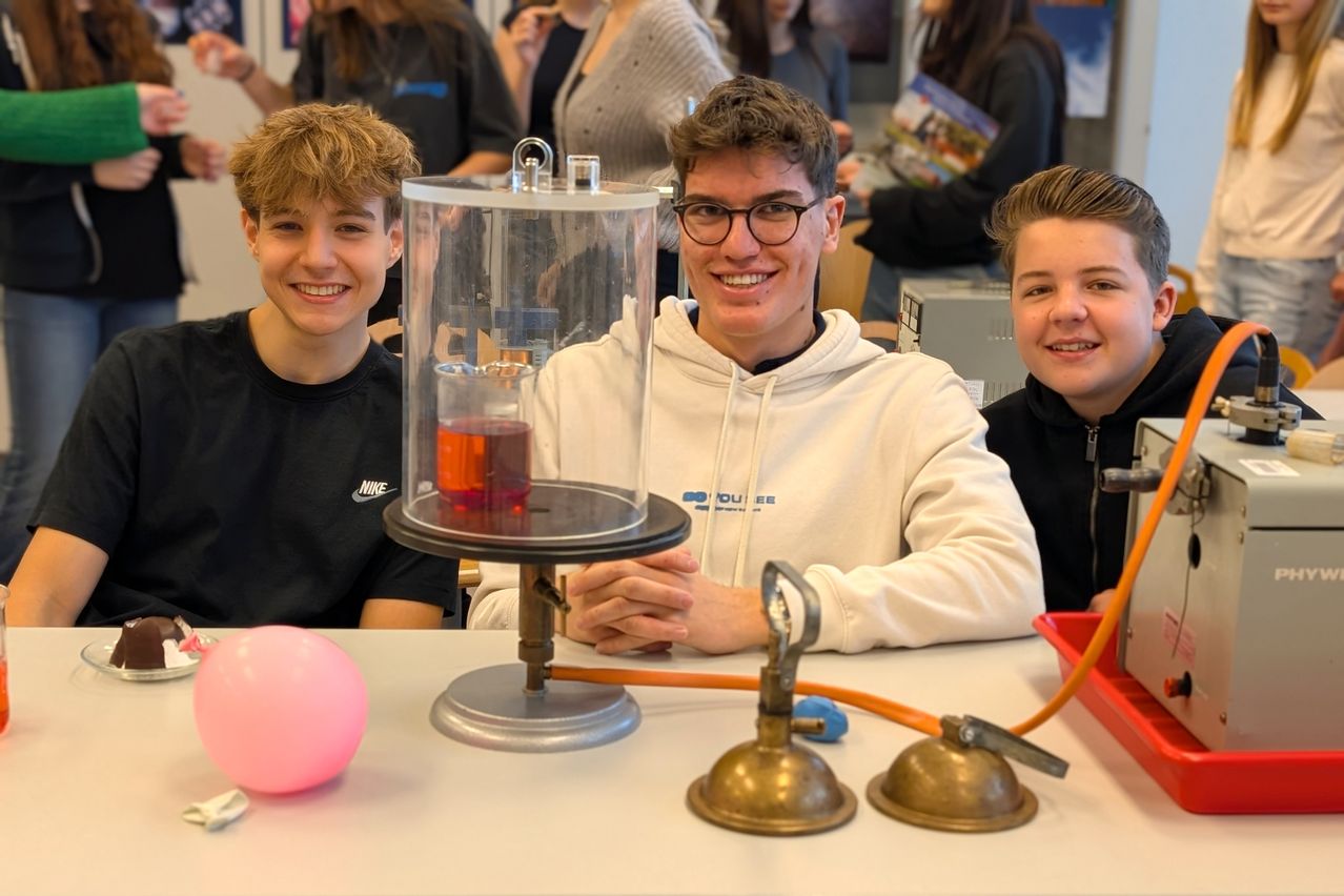 Drei junge Männer in einem Klassenzimmer lächeln, einer in einer weißen Kapuzenjacke mit Brille, der andere in einem schwarzen T-Shirt und der dritte in einer schwarzen Jacke. Sie sitzen um eine Laboreinrichtung herum, die eine rote Flüssigkeit in einem Glasbehälter enthält.