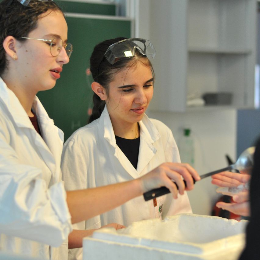 Zwei Mädchen in Laborkittel arbeiten in einem Labor, eine hält ein Messer und schaufelt etwas, während die andere Schutzbrille trägt und zusieht.