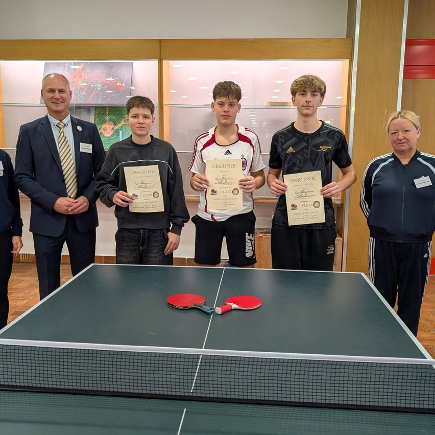 Fünf Personen stehen hinter einem Tisch und halten Urkunden in der Hand, mit zwei Schlägern auf dem Tisch. Sie scheinen eine Leistung zu feiern.