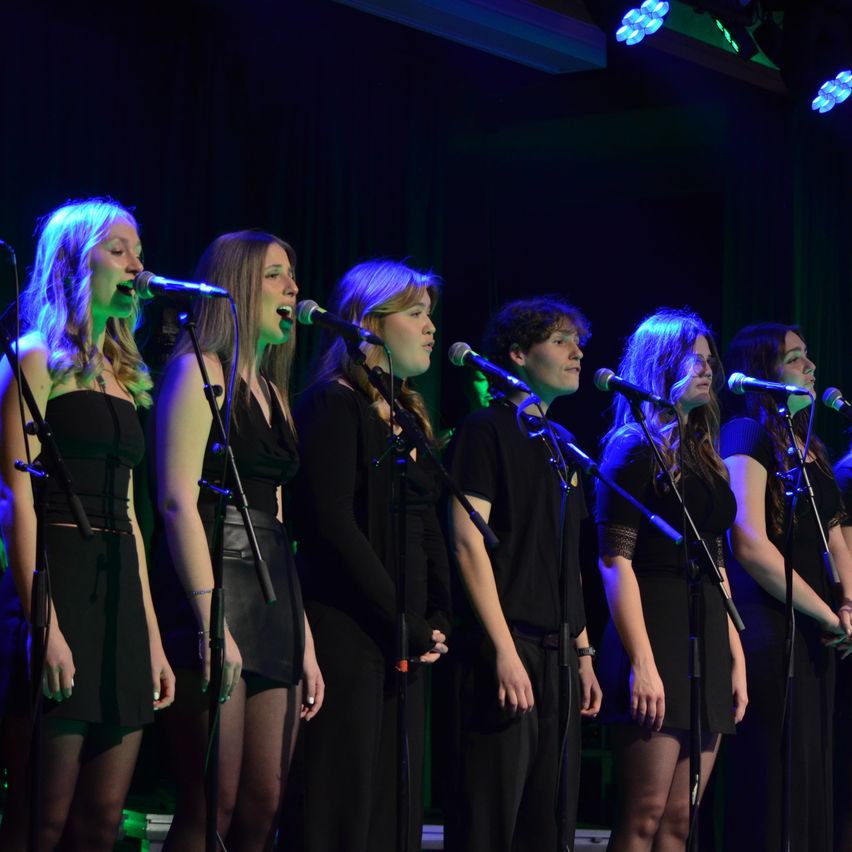 Seven women and a man stand on a stage, singing into microphones. They wear black dresses. The stage is lit with blue and green lights.