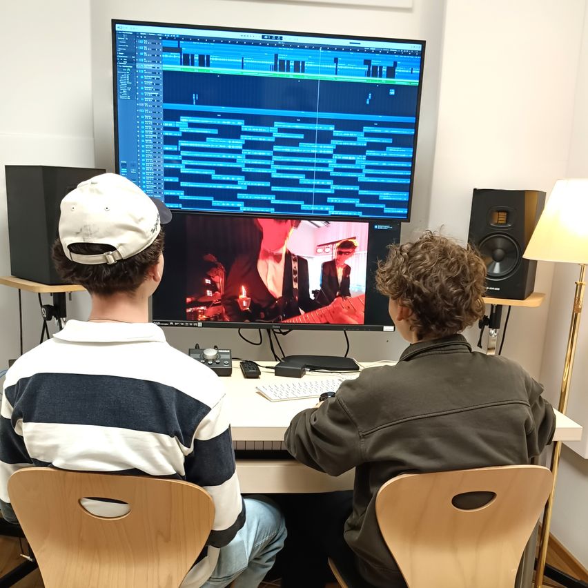 Two individuals sit at a desk, focused on a large monitor displaying an editing software interface. One person wears a baseball cap, while the other is in a green jacket.