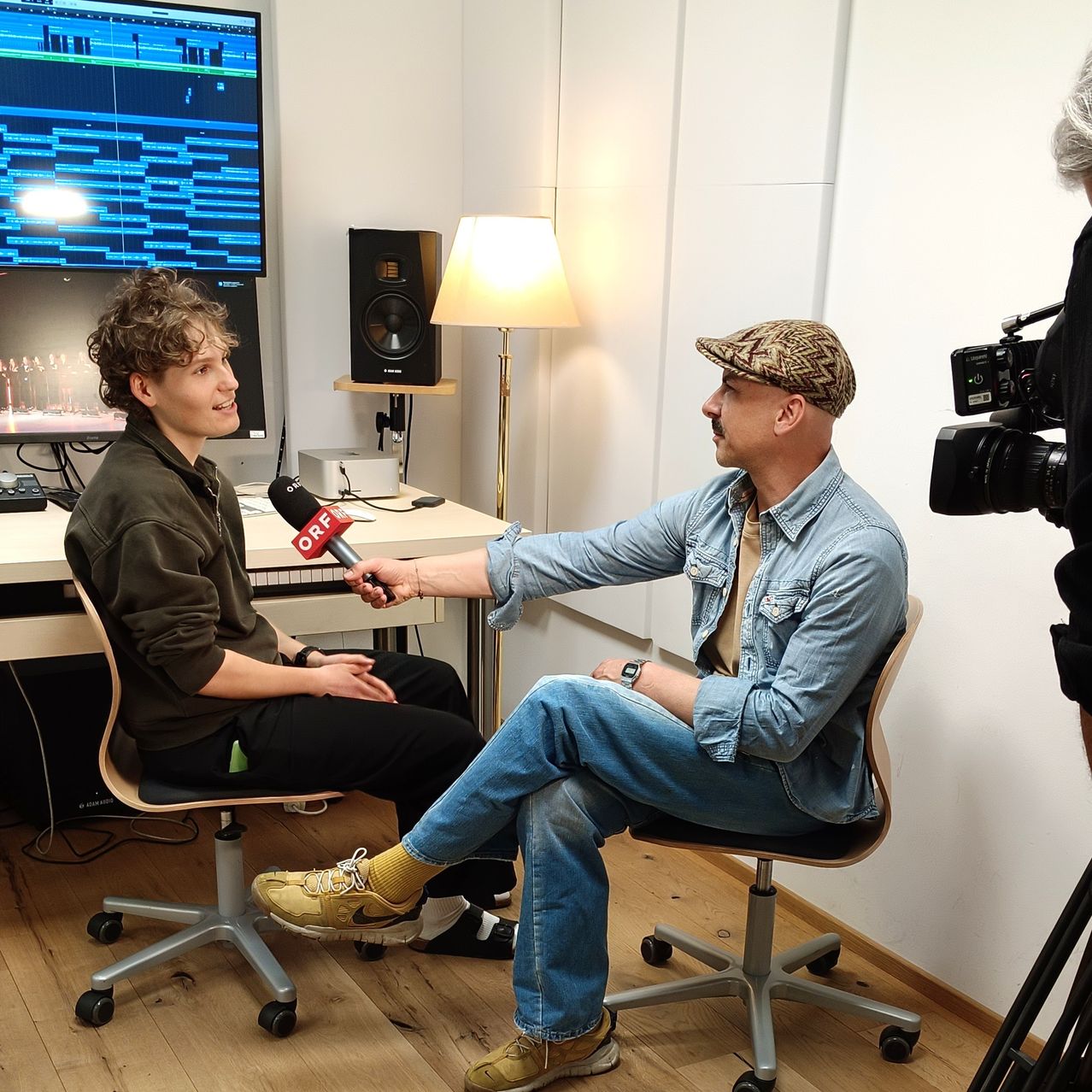 Two men sit in an interview setting, one holding a microphone. The interviewee wears a denim jacket and sneakers. Behind them, a computer and speaker are visible. The interviewer is dressed casually.