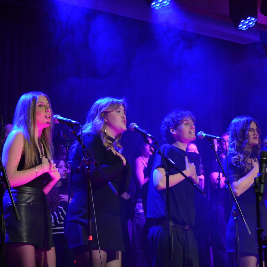 A group of five singers perform on stage under blue stage lighting. The audience watches from behind, and all singers hold microphones.