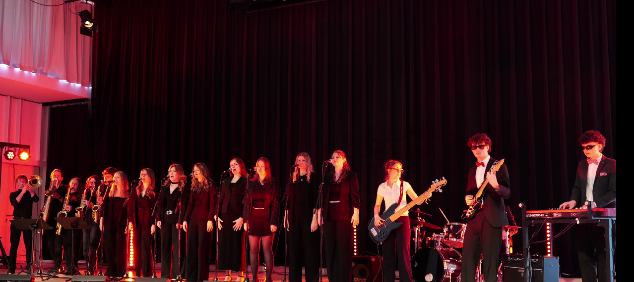 Seven women in black stand on stage with microphones, one plays guitar, another plays drums. Spotlights illuminate the stage.