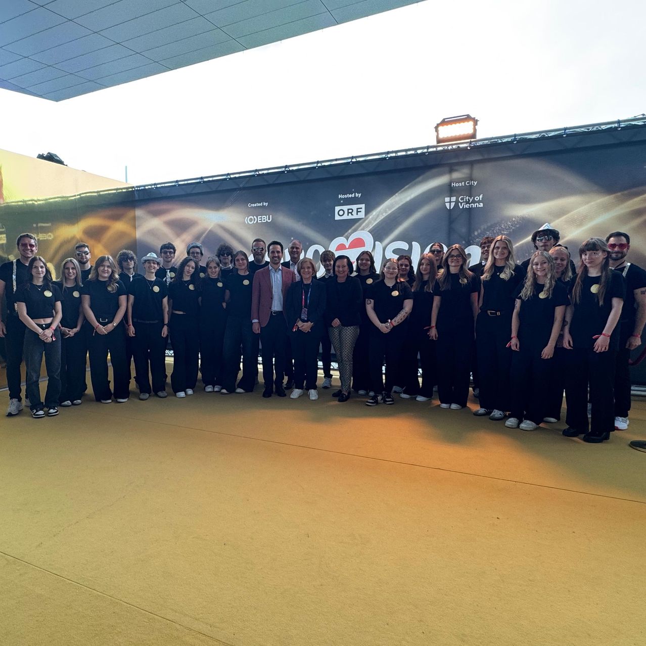 Eine Gruppe von Menschen, die schwarz gekleidet sind, stehen vor einem Hintergrund, einige halten Mikrofone. Sie scheinen für ein Foto zu posieren.