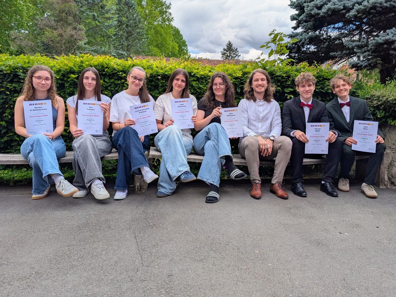 Sieben Schüler, auf einer Bank sitzend, posieren mit Zertifikaten in der Hand. Sie sind lässig gekleidet, manche tragen Turnschuhe, andere Lederschuhe. Hinter ihnen steht ein Strauch und ein Gebäude.