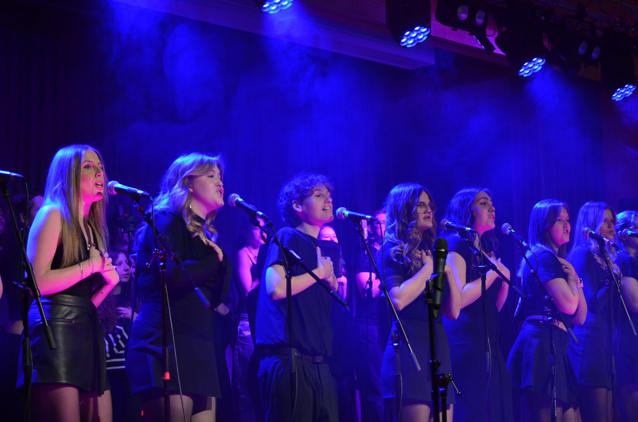 A group of people, likely a choir, performs on stage with microphones under blue lighting, while an audience watches from behind.