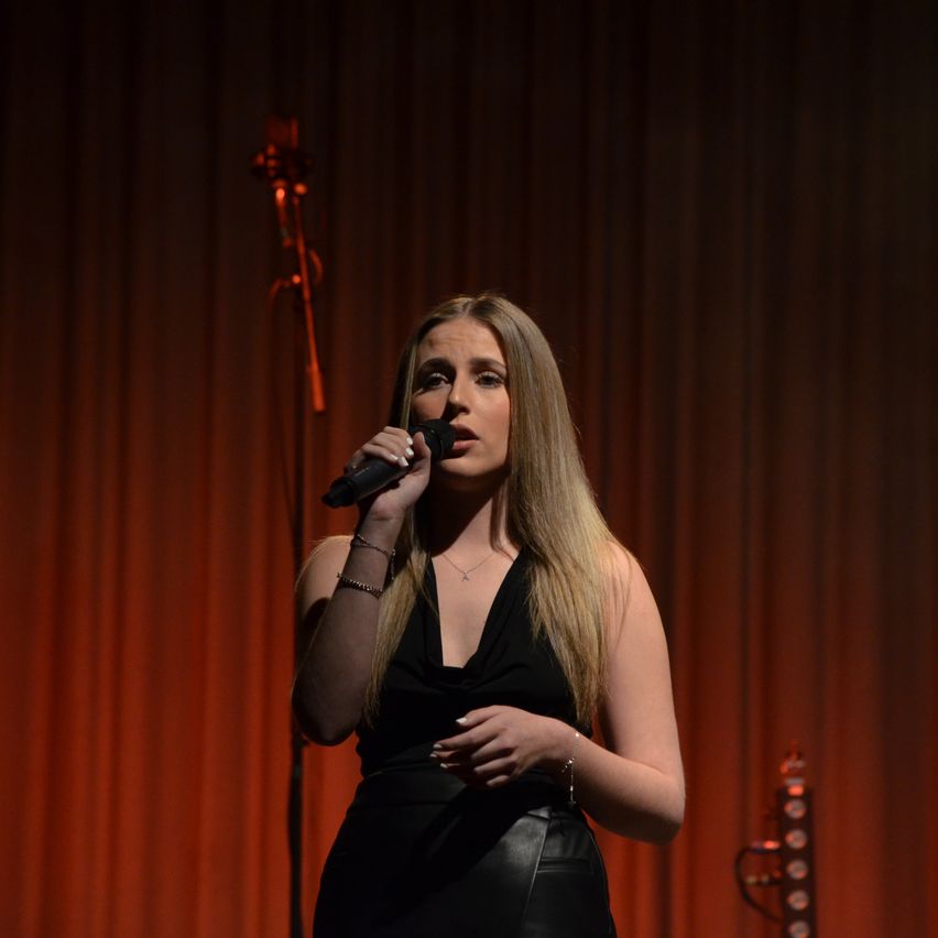 A woman with blonde hair sings into a microphone on a stage with red curtains. She wears a black dress and has jewelry on her wrists.