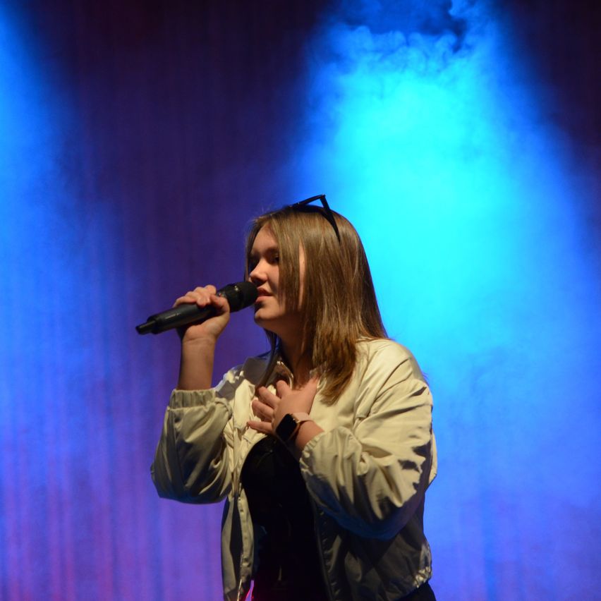 A woman wearing a white jacket and a watch sings into a microphone on stage with blue lighting.