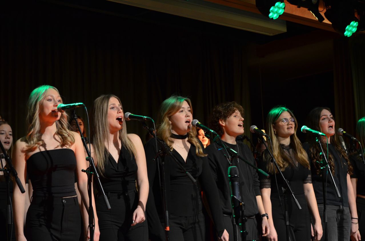 Five women wearing black are on stage singing into microphones. The audience and stage lights are visible in the background.