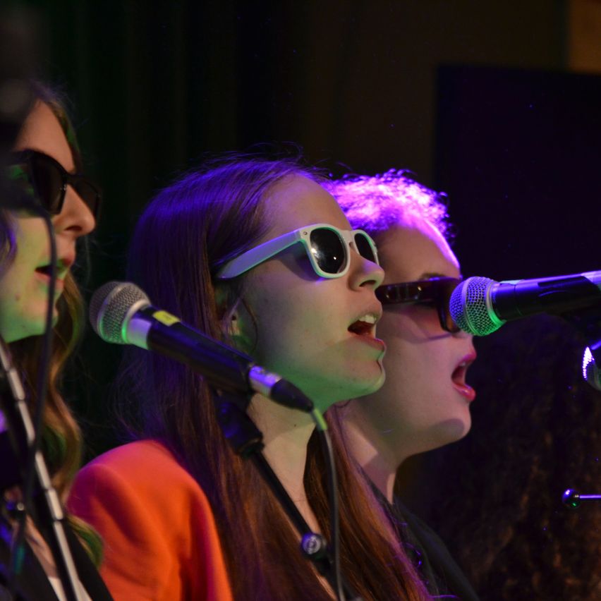 Three women in white sunglasses are singing into microphones at a performance, with one woman wearing an orange top.