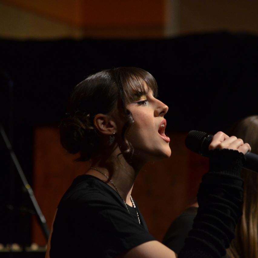 A woman sings passionately into a microphone, her mouth open and eyes closed, with a blurred background.
