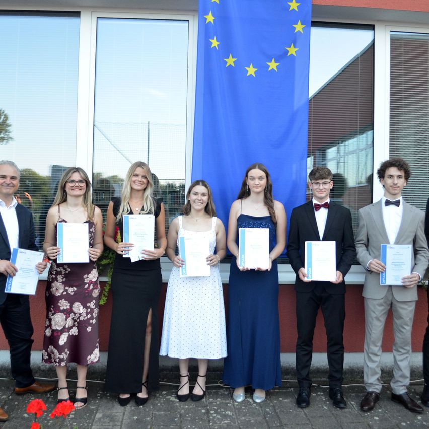 Eine Gruppe junger Erwachsener, sowohl Männer als auch Frauen, posiert für ein Foto und hält Urkunden vor einer Europaflagge.