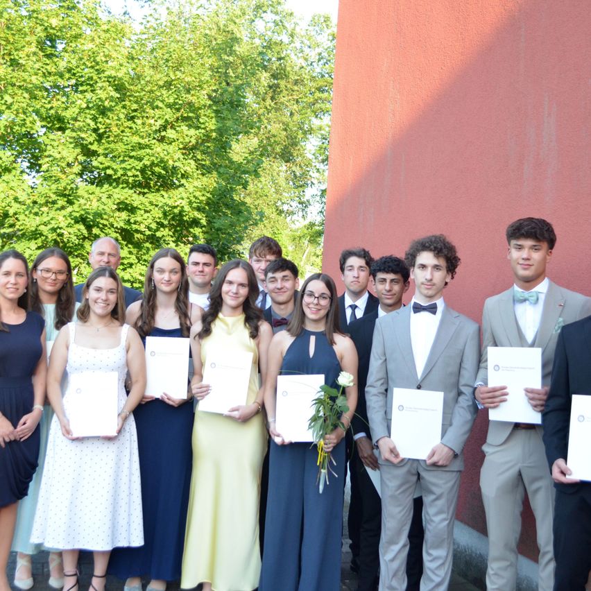 Eine Gruppe junger Menschen steht zusammen mit Urkunden in der Hand, mit einer roten Wand und Bäumen im Hintergrund.