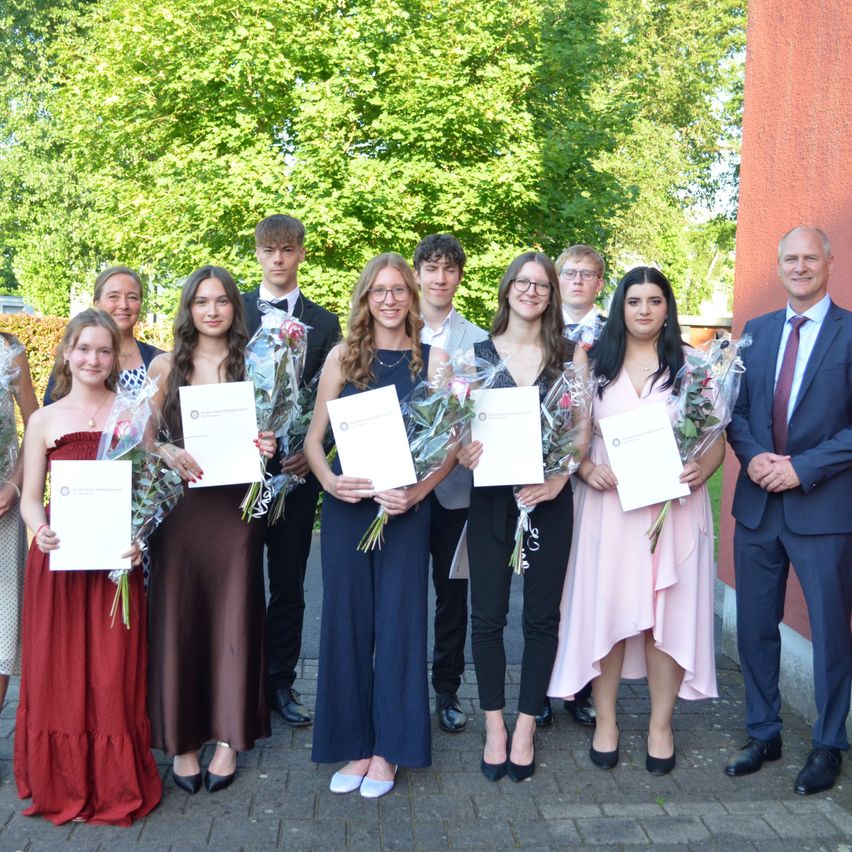 Eine Gruppe junger Menschen und Erwachsene posiert mit Zertifikaten und Blumen in einem Garten draußen für ein Foto.
