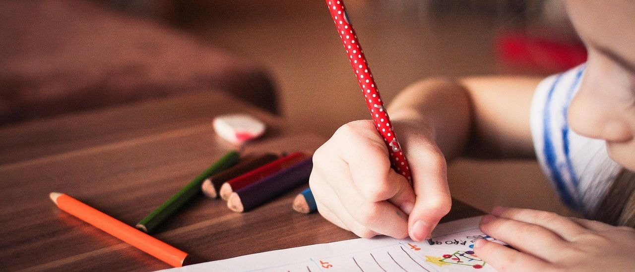 Ein Kind schreibt mit einem roten Stift auf einem linierten Papier. Verschiedene farbige Bleistifte und ein weißer Radiergummi liegen auf dem Tisch.