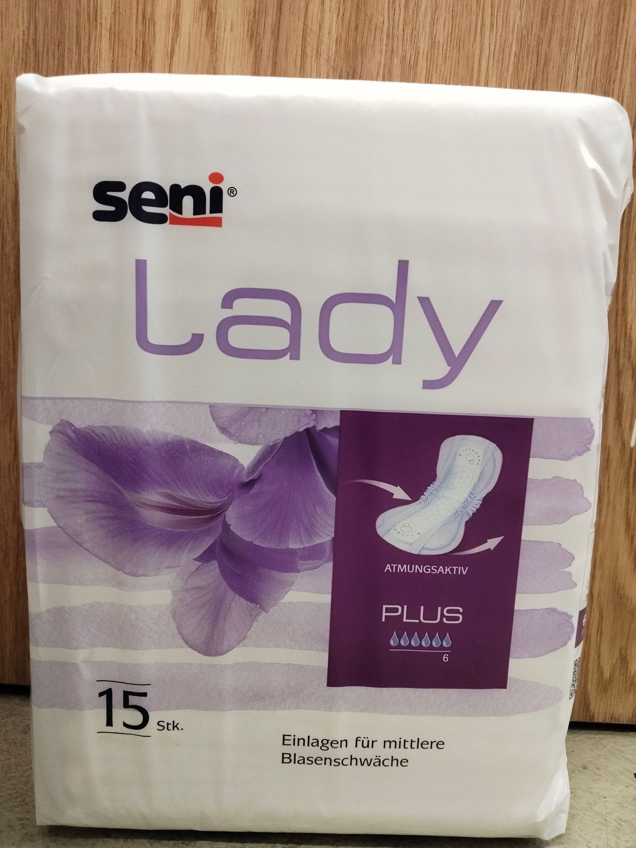 Eine Packung 'seni lady' Damenbinden, gekennzeichnet mit 'Plus' und 6 Stück, enthält 15 Binden. Das Design zeigt eine lila Blume und eine Beschreibung der Atmungsaktivität.