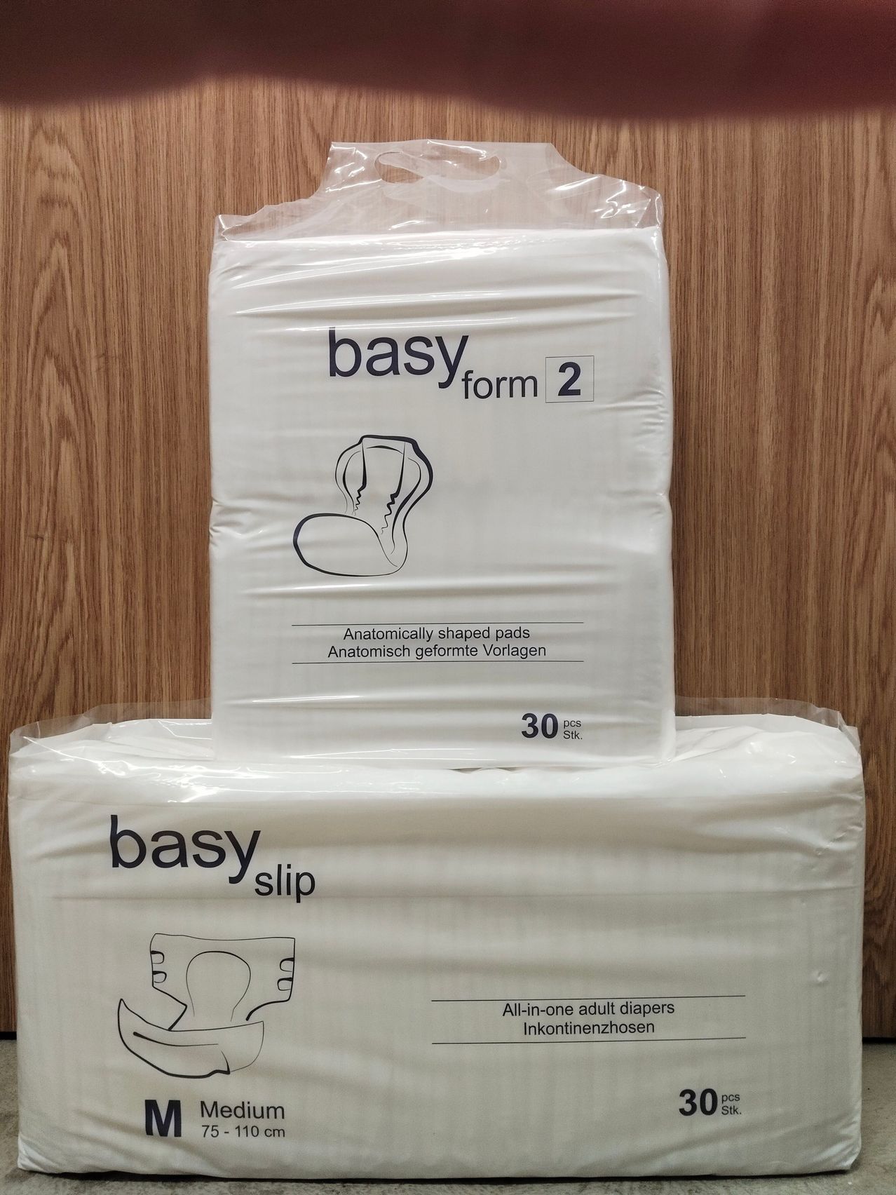 Zwei Packungen Basy-Erwachsenenwindeln. Eine Packung ist mit Form 2 und 30 Stück gekennzeichnet, die andere mit Slip und All-in-One-Design. Beide Packungen sind in Plastik eingewickelt.