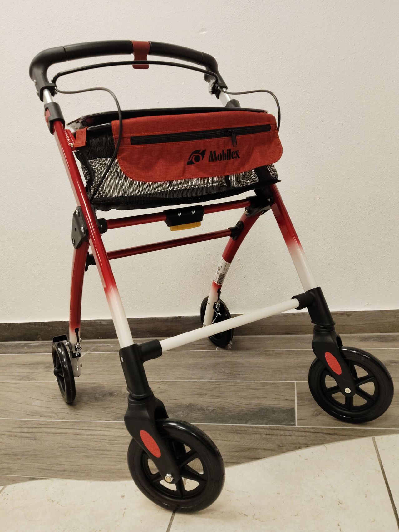 A red and white walker with a red pouch attached to the handle, black wheels, and a label reading 'Mobilex' on the handle. The walker is on a wooden floor.