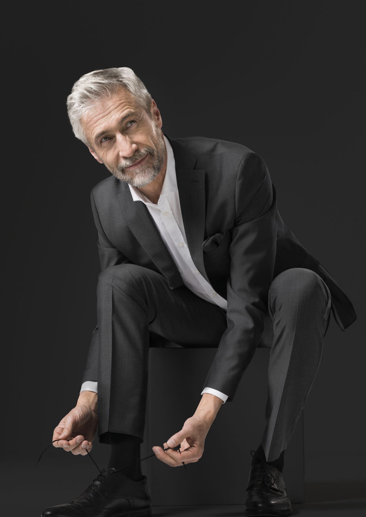 Ein Mann in einem schwarzen Anzug und einem weißen Hemd sitzt auf einer schwarzen Oberfläche, mit einem leichten Lächeln im Gesicht. Er hat graues Haar und einen Bart.