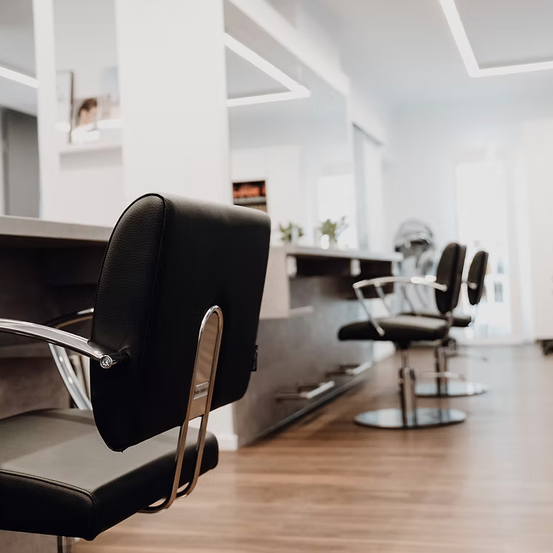 Ein leerer Salon mit schwarzen Stühlen und einem Holzboden. Im Hintergrund sind einige Stühle aufgereiht, und ein Regal mit Pflanzen ist zu sehen.