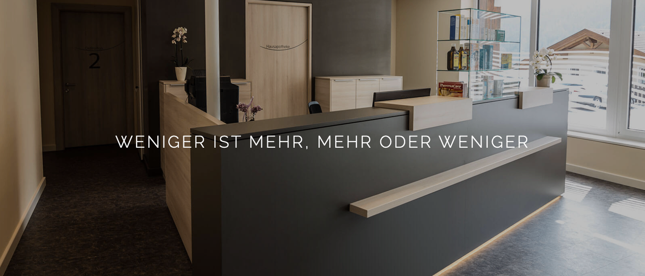Eine Rezeption mit schwarzer Wand und Holztheke. Auf dem Schreibtisch befinden sich zwei weiße Schränke, ein Topf mit Blumen und ein Monitor. An der Wand ist eine Holztür. Es gibt ein Glasregal mit Gegenständen.