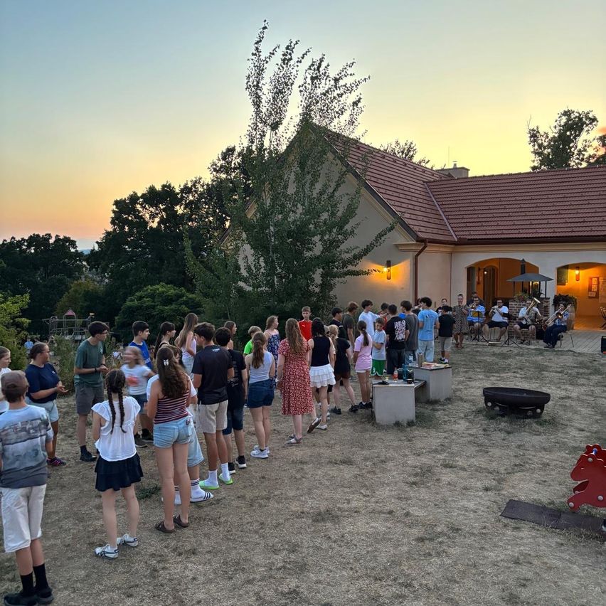 Eine Gruppe von Menschen steht in einer Reihe vor einem Haus mit rotem Dach bei Sonnenuntergang. Bäume und ein Feuerkorb sind im Vordergrund.