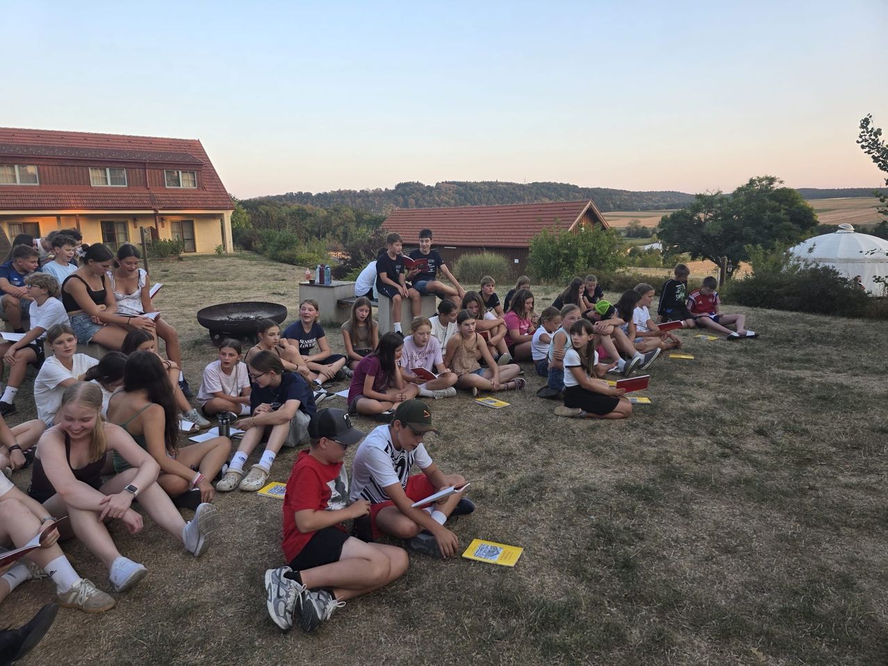Eine Gruppe von Kindern und Jugendlichen sitzt auf der Wiese und liest Bücher und studiert im Freien. Einige sitzen auf dem Boden, während andere auf einer Betonbank sitzen. Die Umgebung umfasst ein Haus, Bäume und ein Feuerloch.