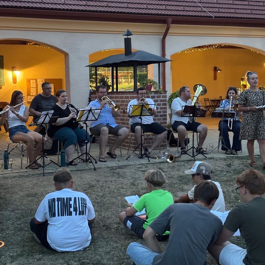 Eine Gruppe von Musikern, darunter Trompeten, Posaunen und Flöten, spielt auf einer Bühne vor einem Publikum. Die Musiker sitzen auf Stühlen mit Notenständern, und einige stehen. Das Publikum sitzt auf dem Gras.