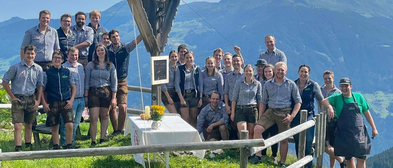 Eine Gruppe von Menschen, gekleidet in traditioneller bayerischer Tracht, posiert für ein Foto auf einem Berg mit einer Holzstruktur im Hintergrund. Die Umgebung umfasst einen Tisch mit Blumen und einer Kerze.